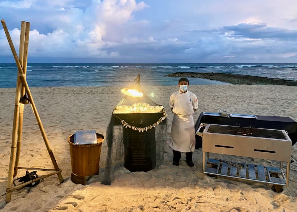 A chef on stands next to a grill on the beach.