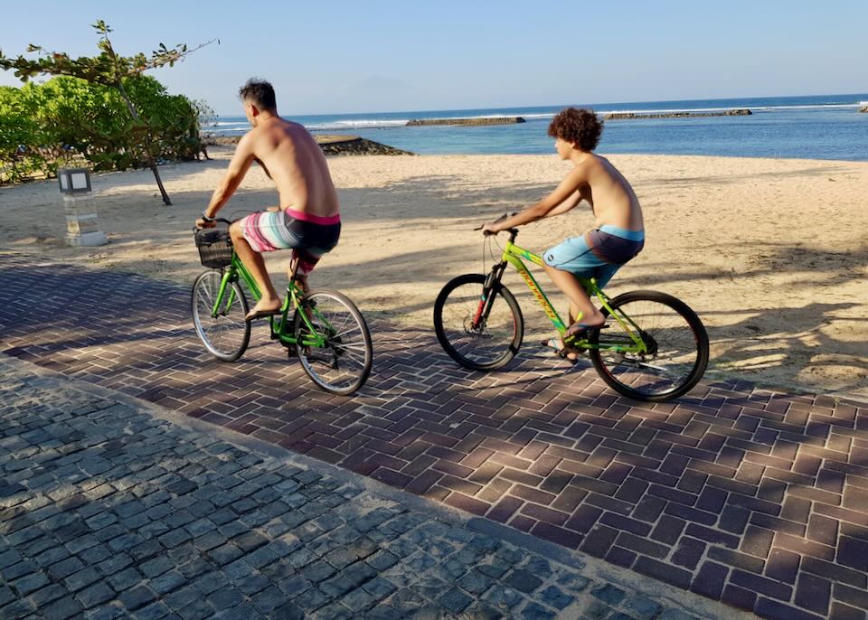 Two men ride bikes on a path at the beach.