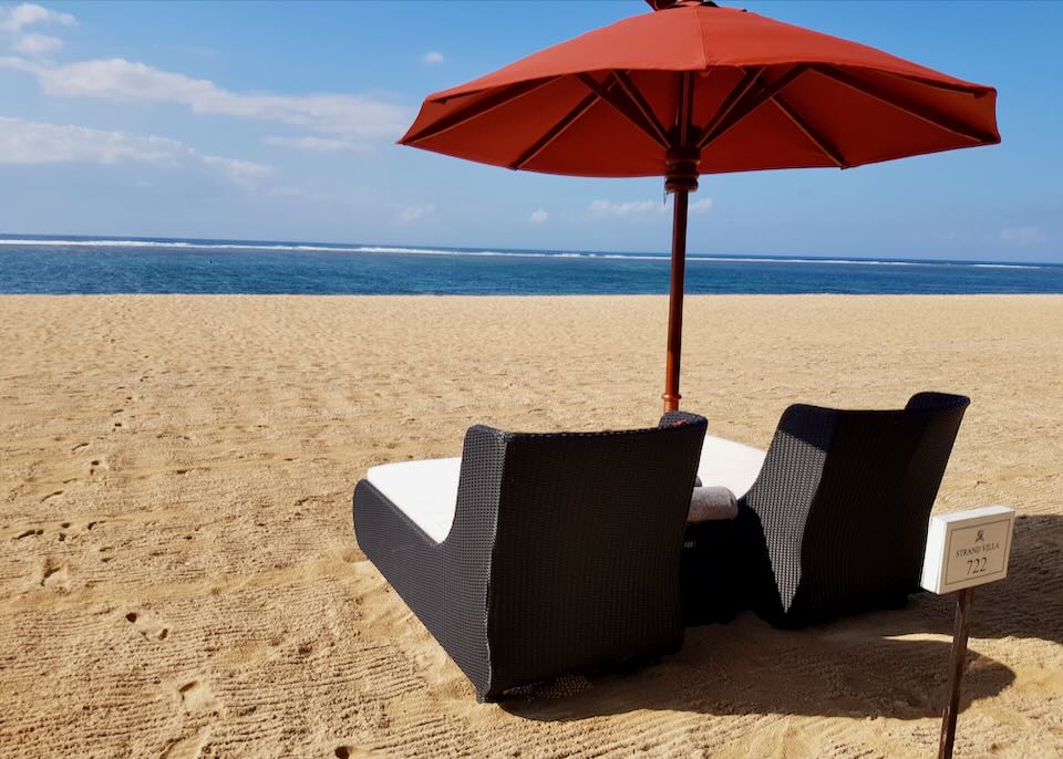 Two beach-front chairs sit under an orange umbrella with a reserve sign for villa 722.