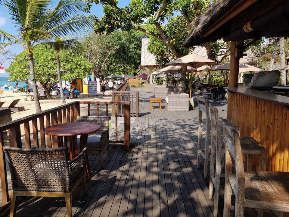A wood deck with wicker and wood tables and chairs sits by the beach.