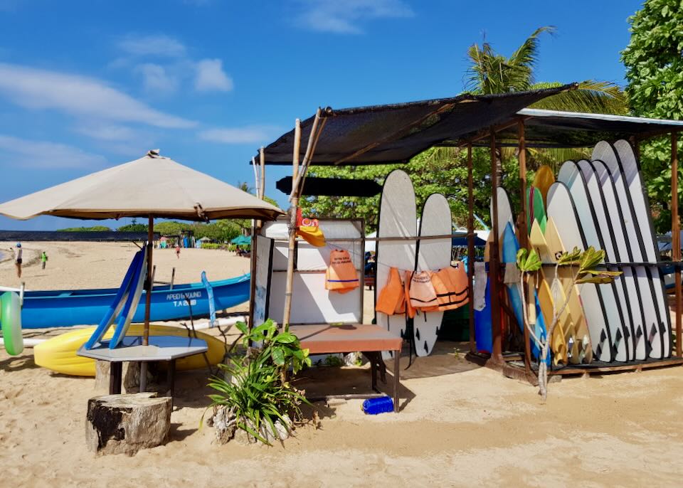 Paddle boards, canoes, and life jackets sit under a canopy on the beach.