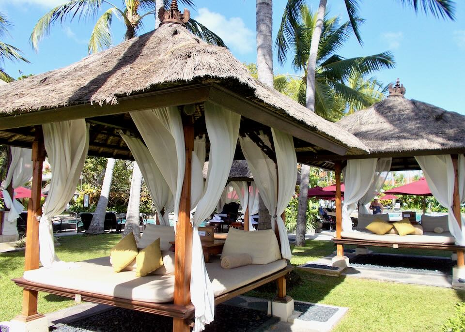 Thatched-roof gazebo lounges with mattresses, pillows, and flowing curtains sit on the lawn.