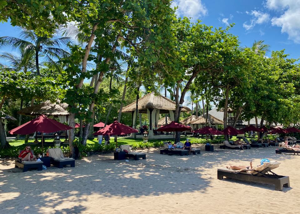 Facing away from the water, lounge chairs with red umbrellas ate shaded under tall trees.
