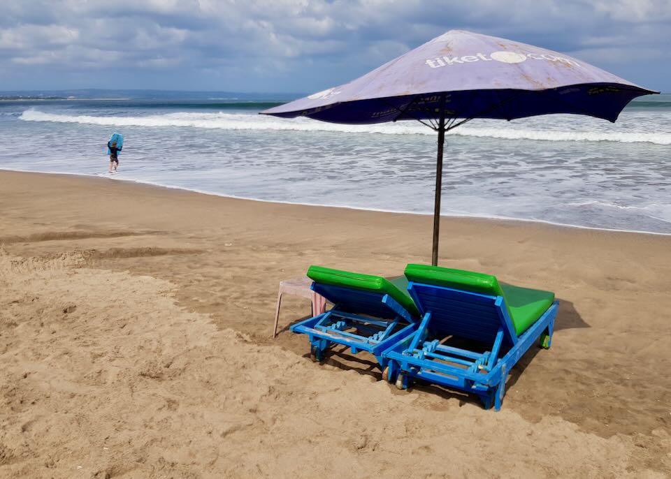 Two lounge chairs sit under an umbrella by the ocean.