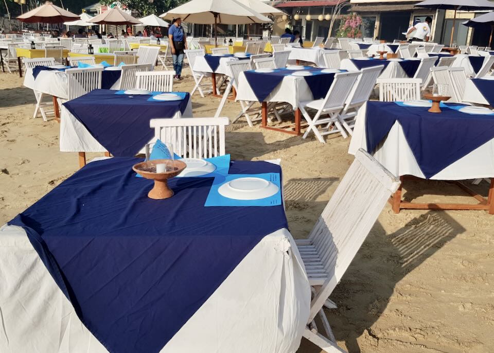 White tables with blue tablecloths sit on the beach.