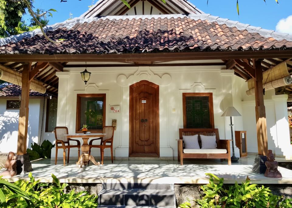A white painted bungalow with a thatched roof with wood doors and plants surrounding it sit in the sun.