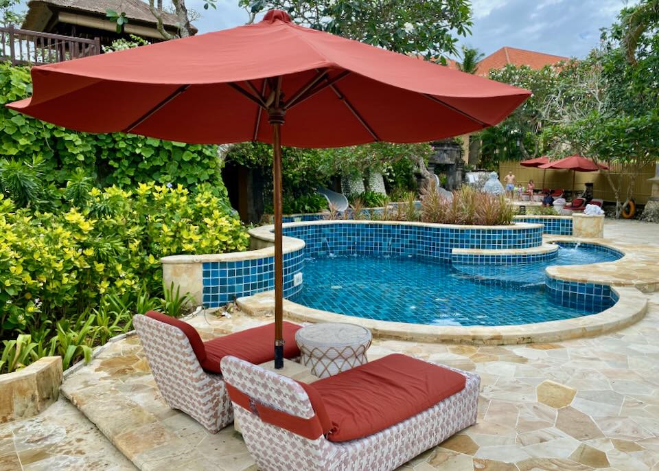 A small shallow pool is next to bright orange lounge chairs under an umbrella.
