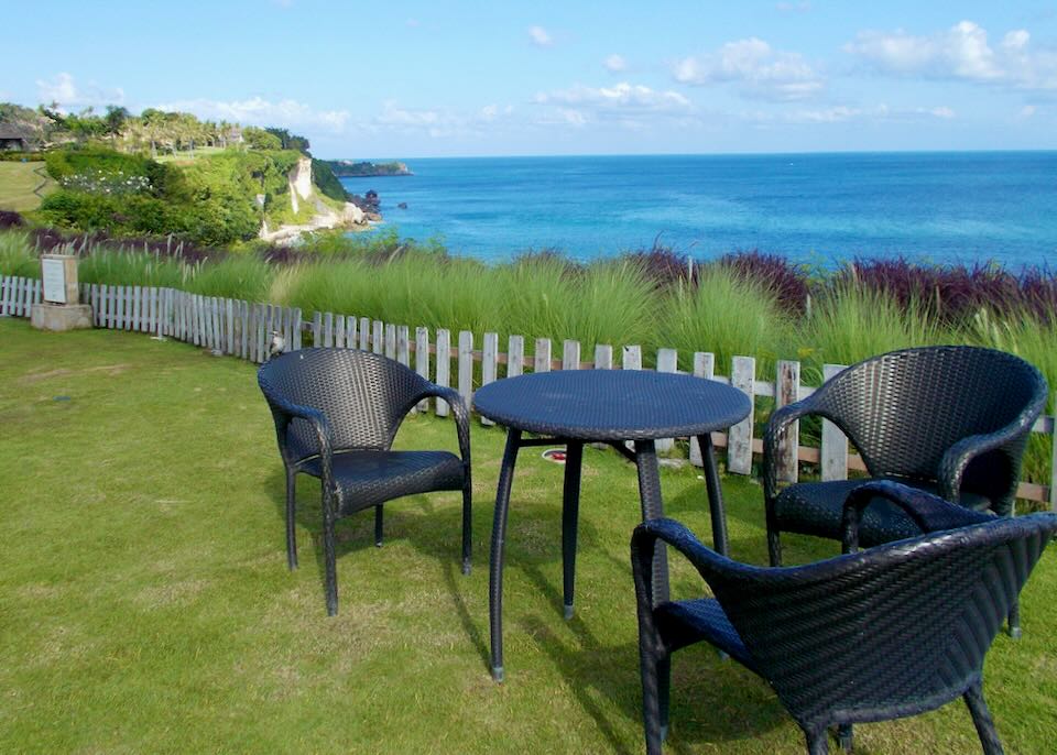A table and chairs sits next to tall grass and a fence overlooking the ocean.