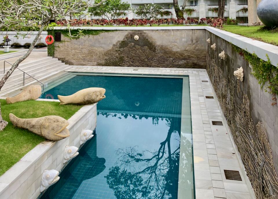 A small "L" shaped pool with stone fish sculptures sits below a retain wall of fountain flowers.