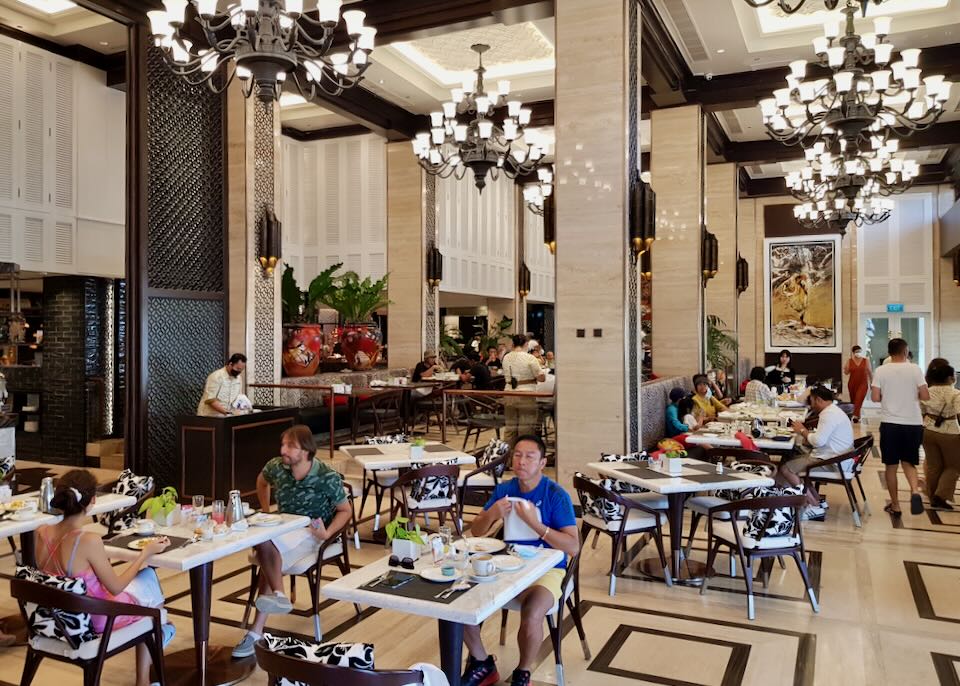 People eat breakfast at smaller tables in a room with high ceilings and large black and white chandeliers.