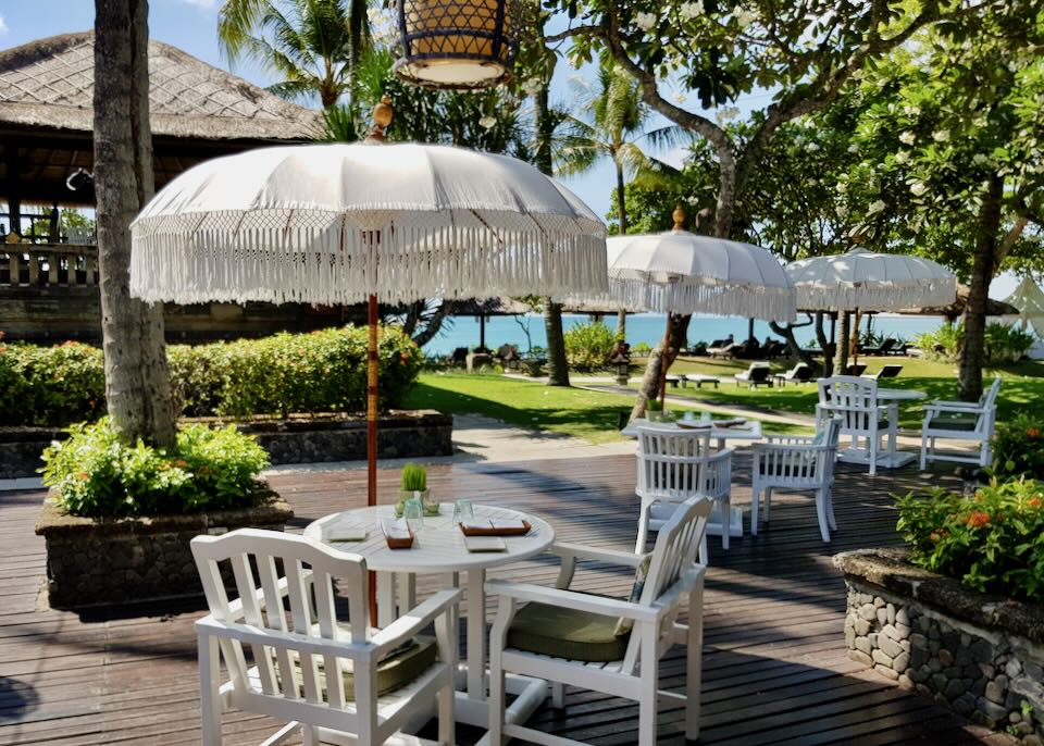 White tables with white umbrellas and fringe sit outside on a brown wood patio overlooking the ocean.
