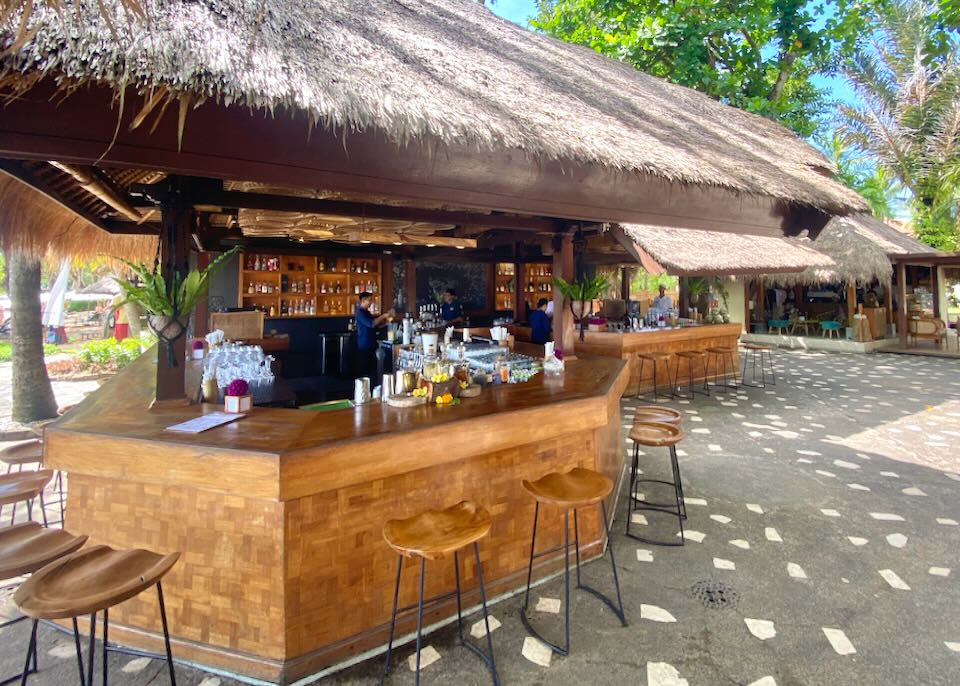 Bartenders work behind a wooden outdoor thatched-roof bar.