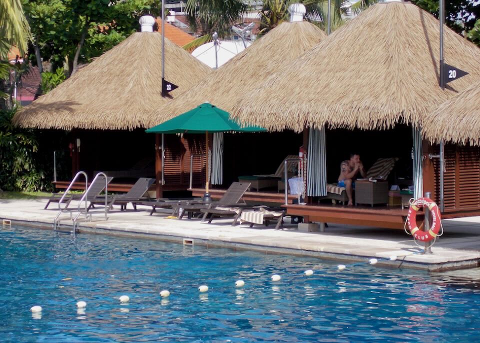 A couple sit under a thatched-roof cabana by the pool.