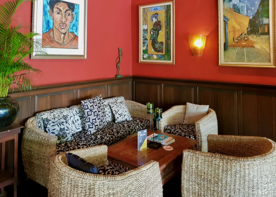 A cozy corner filled with a whicker loveseat, chairs, and a table with colorful art on a red wall.