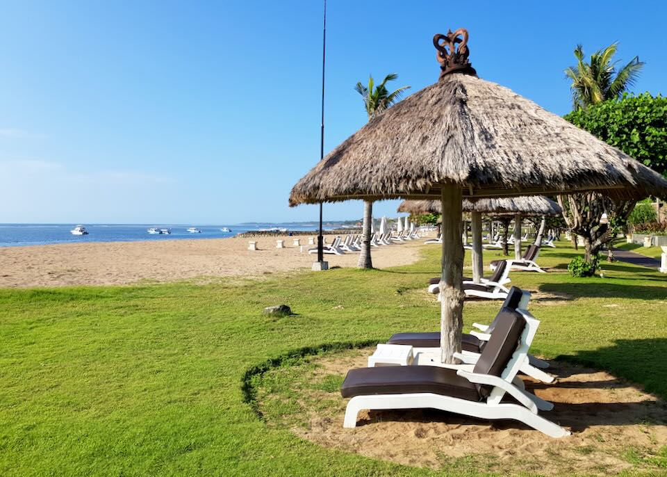 Cushioned lounge chairs sit under thatched-roof canopies on the green lawn next to the beach.