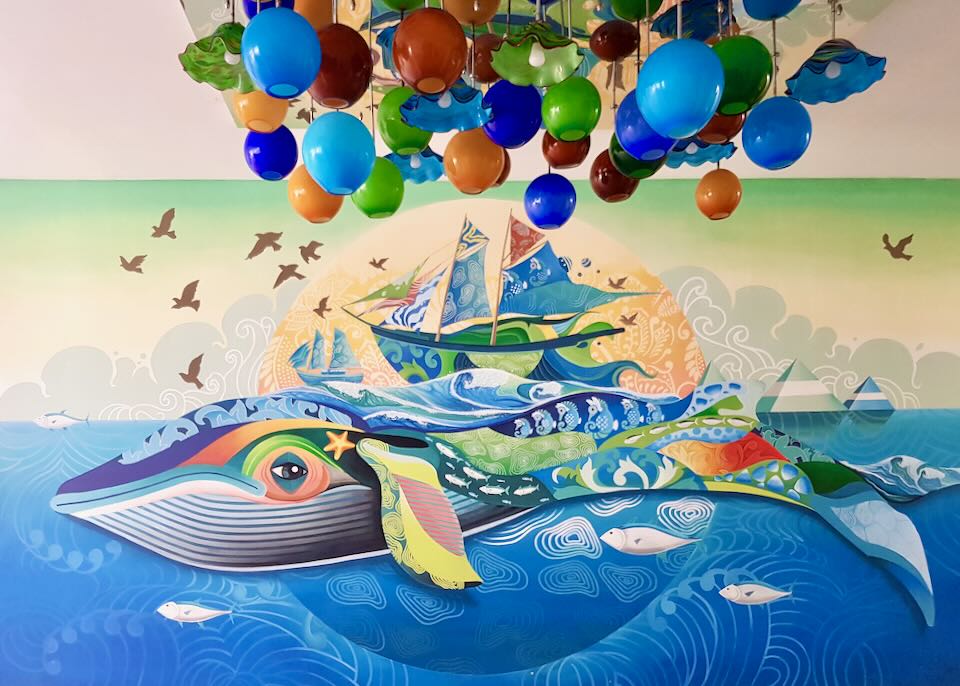 A colorful mural of a whale and boat sit under a colorful blown-glass light fixture.