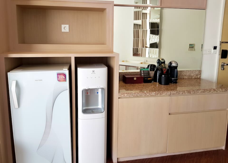 A small refrigerator, water cooler, and coffee makers sit in the hotel room.