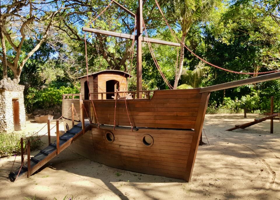 A wood playground boat sits in the sand with steps leading to its deck.