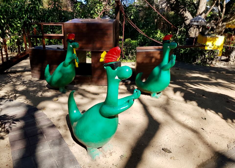 Three sculptures of green dinosaurs sit in a sand pit by a playground boat.
