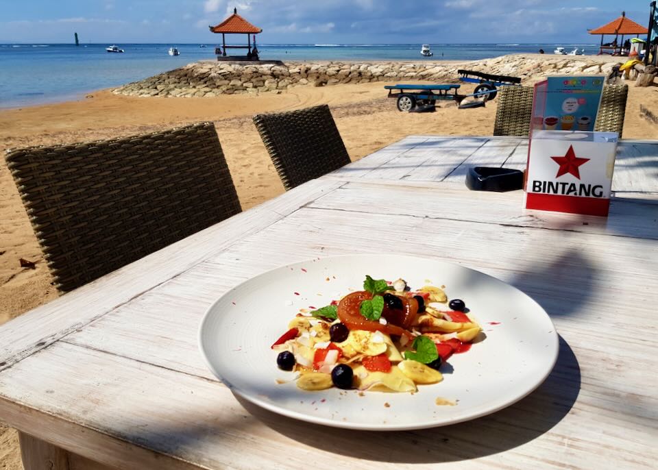 A plate of crepes with berries sits on a wood table on the beach.