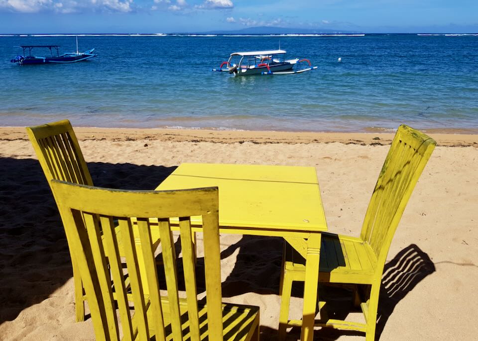 A yellow painted wood table and chairs sits on the sand with views of fishing boats near the shore.
