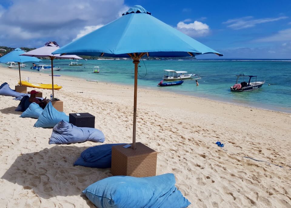 Blue bean bags sit under umbrellas on the beach.