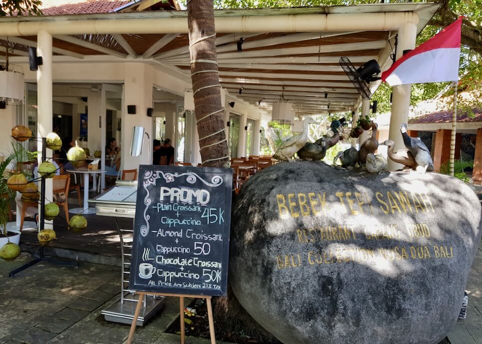 A large rock sits outside a restaurant with the name Bebek Tepi Sawah carved into it.